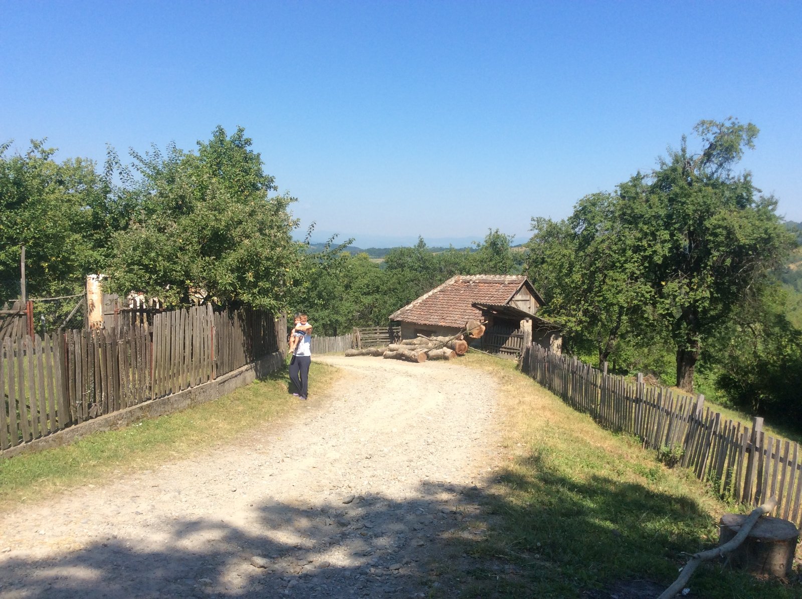 A track through Planina village, with a man and small child walking, wooden fences either side and rural buildings beyond